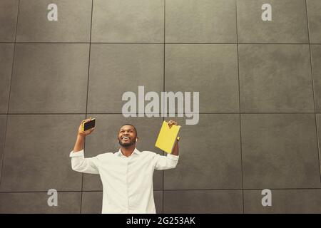 Joyful black businessman staying, looking up, making call. African businessman holding yellow laptop and smartphone, received great news read email Stock Photo