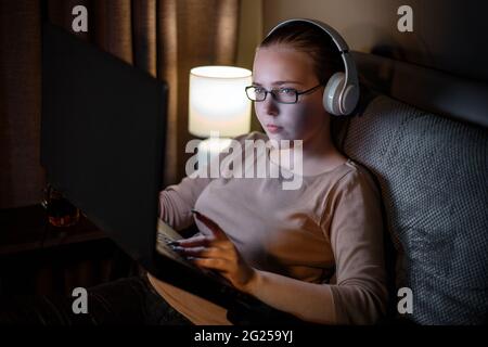 Young woman in glasses studying or working using laptop at night in home interior. Overtime sofa workspace. Attentive serious teenager student studies Stock Photo