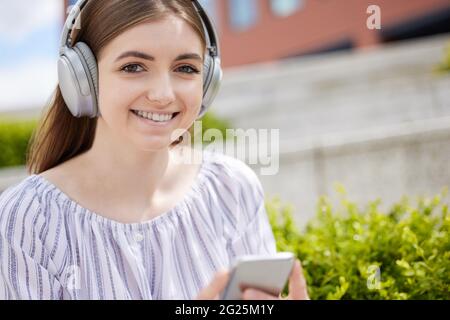 Portrait Of Smiling Young Woman Student On College Campus Streams Music From Mobile Phone To Wireless Headphones Stock Photo