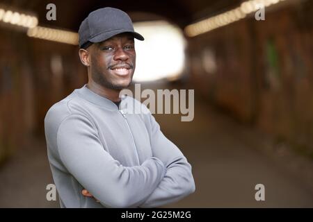 Portrait Of Smiling Young Man Wearing Baseball Cap Standing In Tunnel In In Urban Setting Stock Photo