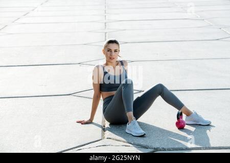 Portrait of a beutiful brunette resting from exercising with little kettlebell. She is sitting on the pavement outdoors. In grey top and yoga pants. O Stock Photo