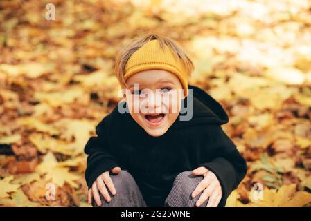 Cute kid, boy 5 years old in black hoodie and yellow crown having fun at autumn street, jumping and running around on carpet of fallen leaves Stock Photo