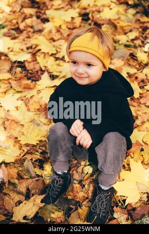 Cute kid, boy 5 years old in black hoodie and yellow crown having fun at autumn street, jumping and running around on carpet of fallen leaves Stock Photo