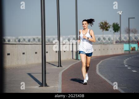 Happy immersed athletic woman running on the new track outdoors, listening to the music. On a sunny day under a clear blue sky. She is wearing white s Stock Photo