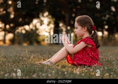 Girl sits on grassy lawn examining clover flower in evening sun Stock Photo