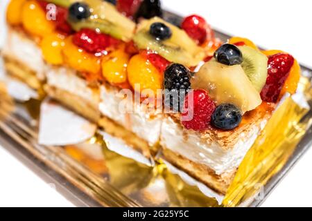 close up of a puff pastry and cream cake with red wild berries, kiwi fruit pieces, pineapple, mandarin slices on a white table, selective focus on one Stock Photo