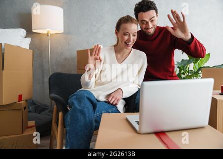 Smiling couple with laptop sitting on floor in new house, waving hands Stock Photo