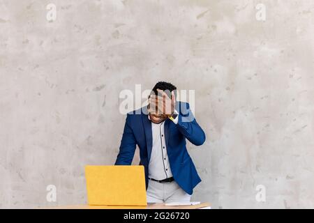 Stressed and tired African American man standing in office or at home leaned over yellow laptop holding online meeting, texting client, student learn Stock Photo