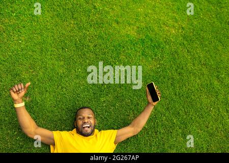 Top view of cheerful overjoyed African American male celebrating victory, successful race, game or football match. Young afro laying on the grass Stock Photo