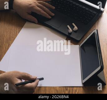 several sheets of paper on the office table while a man uses a laptop computer with small headphones and a tablet to make video conferences about new Stock Photo