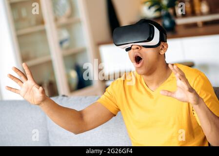 Excited latino carefree guy, sitting on a sofa at home and gesturing hands while testing new app via VR device. Cheerful modern hispanic man using VR glasses, virtual reality concept Stock Photo