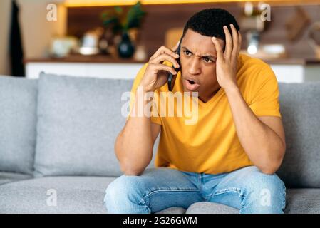 Confused hispanic guy sitting on the couch in the living room, dressed in casual shirt, bewildered, talking on the phone, angry, annoyed, have unpleasant telephone conversation Stock Photo