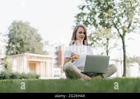Portrait of young adult Woman texting laptop, sitting on the grass outside in park. Working and drinking lemonade soda. Using laptop. Distance learnin Stock Photo