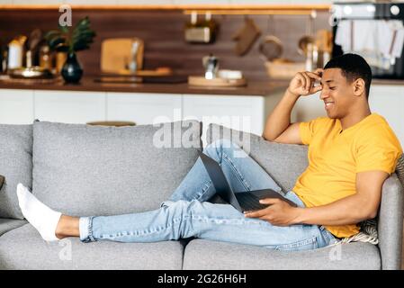 Positive attractive hispanic guy sitting on a sofa at home in the living room, using a laptop, browsing the Internet, chatting online on social networks, looking at laptop, smiling Stock Photo