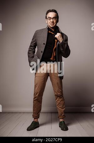 Optimistic stylish adult man hipster in pants and plaid jacket stands holding hand in pocket and taking off his scarf Stock Photo