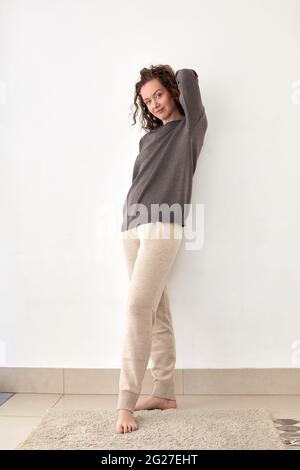 Full body of young barefoot female standing with hand behind head and smiling at camera Stock Photo