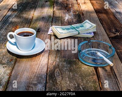 Cup of coffee with an ashtray and money on a wooden table. Coffee drink. A cigarette in an ashtray. A bundle of money. American dollar. Wooden table. Stock Photo