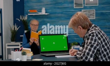 Senior woman looking at green screen monitor working from home, taking notes on notebook. Elderly businesswoman watching desktop laptop display with green mockup, chroma key, while man reading a book Stock Photo