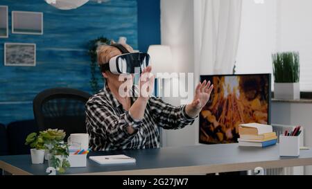 Retired woman experiencing virtual reality using vr headset in living room working from home. Old remote employee searching, analysing financial reports while elderly wife watching tv in background Stock Photo