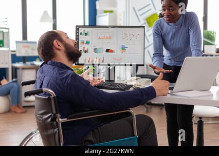 African bookkeeper working with handicapped invalid immobilized colleague calculating profit on laptop, taking notes on pc, analysing financial documents sitting at desk in start-up financial company. Stock Photo
