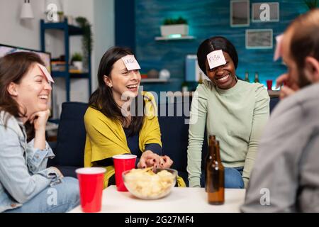 Mixed race friends gathering in living room playing guess who game using sticky notes. Mixed race people having fun, laughing together while sitting on sofa late at night during funny party. Stock Photo