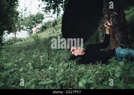 Young handsome man in the forest holding a black umbrella and lying on the grass Stock Photo