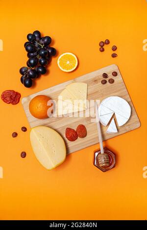 Wooden board with different cheeses, oranges, nuts, dried apricots, grapes and honey. Top view, flat lay. Orange background. Stock Photo
