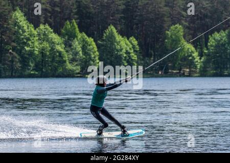 Girl with helmet and life vest rides a wakeboard on the lake with forest on background, wakeboarding, water sport in summer Stock Photo