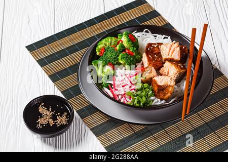 Salmon teriyaki with rice noodle, broccoli, radish and green onion in a bowl with chopsticks on a white wooden table on a bamboo mat Stock Photo