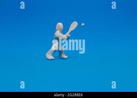 White plasticine doll practicing tennis on a blue background. The doll holds the racket ready to hit the ball coming towards him. Stock Photo