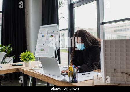 african american plus size engineer in medical mask using laptop near blueprint and house model in office Stock Photo