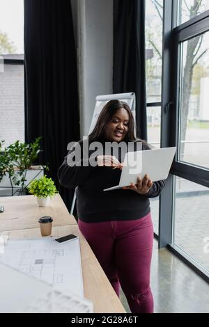 smiling african american plus size engineer holding laptop near blueprint on desk Stock Photo