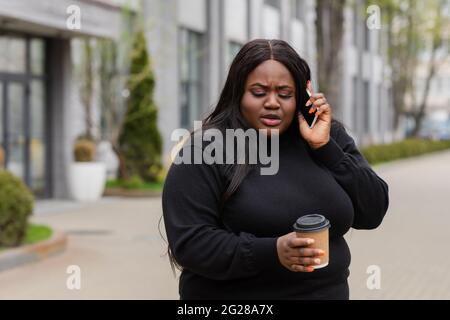 african american plus size woman holding coffee to go and talking on smartphone outside Stock Photo