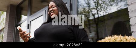 low angle view of cheerful african american plus size woman holding coffee to go and looking at smartphone outside, banner Stock Photo