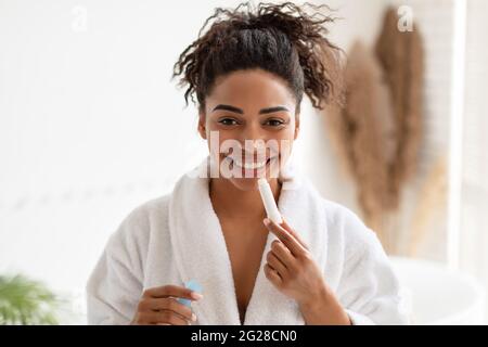 African Lady Applying Lip Balm Smiling To Camera In Bathroom Stock Photo