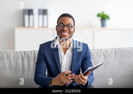 Portrait of happy black male psychologist looking at camera and taking notes during therapy session at clinic Stock Photo