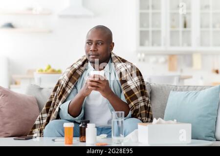 Thoughtful african american man feeling sick, holding mug with hot drink, sitting on sofa near table full of pills Stock Photo