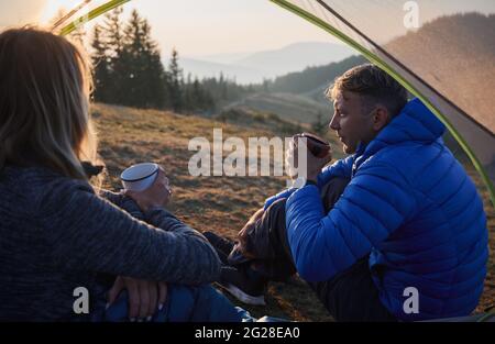 Close up view from inside tent of couple of friends sitting inside tent and drinking hot tea while looking at beautiful environment. Camping in the mountains at sunny day. Stock Photo