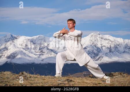 Wushu master in a white sports uniform training on the hill. Kungfu champion trains maritial arts in nature on background of snowy mountains. Stock Photo