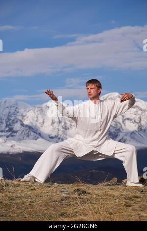 Wushu master in a white sports uniform training on the hill. Kungfu champion trains maritial arts in nature on background of snowy mountains. Stock Photo