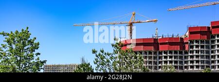 Construction cranes over the houses with red roofs against a blue sky with green leaves of trees in the foreground. Construction concept. Banner forma Stock Photo