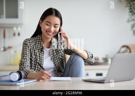 Smiling asian woman freelancer talking on phone and taking notes Stock Photo