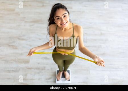 Indian female measuring waist while standing on scales, happy about successful weight loss result at home, above view Stock Photo