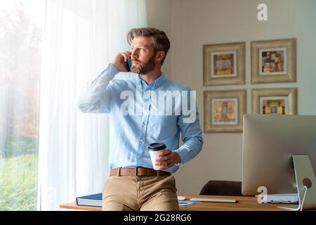 Shot of handsome businessman sitting at desk and talking with someobdy on his mobile phone while working from home. Stock Photo