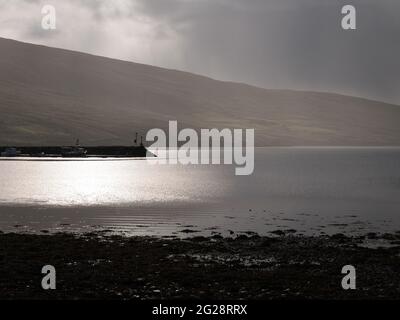 A grainy, moody almost black and white view of sunlight on the the water and the jetty at Voe at the head of Olna Firth on Mainland, Shetland, UK Stock Photo