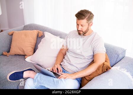 Shot of handsome man looking thoughtfully while sitting on sofa at home and working on laptop. Home office. Stock Photo