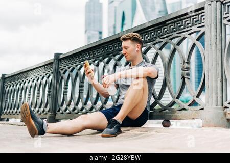Tired Young man runner sitting on road, using mobile phone and relaxing after sport training. Holding water bottle while doing fitness workout in city Stock Photo