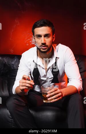 Bearded man with glass of whiskey smoking cigar in armchair indoors ...