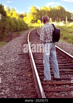 Elderly man in plaid shirt and denim pants with black backpack stands on rails of railway track. Stock Photo