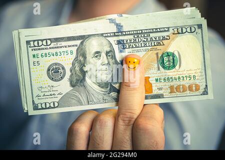 Happy young man with dollars. The businessman holds the cash in his hand. A funny smiley face is drawn on the yellow nail. The concept of high income Stock Photo